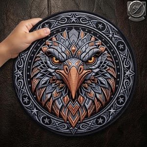 Può includere: Una toppa circolare con un design dettagliato di testa d'aquila in tonalità metalliche argento e rame. L'aquila ha occhi gialli penetranti e un becco affilato, circondata da un bordo di stelle e intricati motivi a intreccio su sfondo nero.