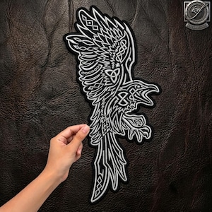 May include: A silver and black embroidered patch featuring a stylized raven with intricate knotwork patterns throughout its wings and body. The design is depicted in a profile view with sharp, angular feathers and a distinct beak, set against a dark background.