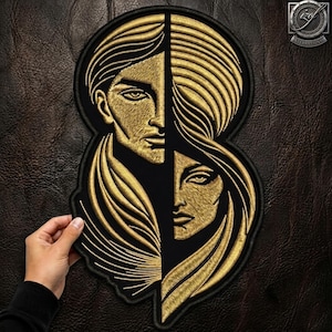 Könnte beinhalten: Ein schwarz-goldener Stickaufnäher mit einem geteilten Gesichtsmotiv. Die linke Seite zeigt ein Gesicht mit kurzem Haar, während die rechte Seite ein Gesicht mit langem, fließendem Haar darstellt. Stickerei mit metallischem Goldgarn auf schwarzem Stoff.