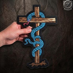 May include: An embroidered patch featuring a brown wooden cross with a textured surface. A bright blue snake with a pink tongue is coiled around the vertical and horizontal beams of the cross. The design is outlined in black thread on a dark background.