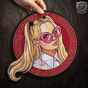 May include: A circular embroidered patch featuring a blonde character with pigtails wearing large pink cat-eye glasses. The character is dressed in a white collared shirt, set against a deep red background with intricate geometric border patterns.