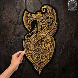 May include: A gold-coloured embroidered patch featuring a detailed battle axe design with intricate Celtic knotwork, a central helmeted figure, and a tree of life motif. The patch has a black border and is shaped to follow the axe's ornate curves.