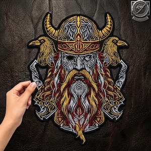 May include: An embroidered patch featuring a central figure wearing a horned helmet and an eyepatch, flanked by two ravens. The design incorporates intricate gold, silver, red, and black thread work, depicting a bearded face with detailed patterns.