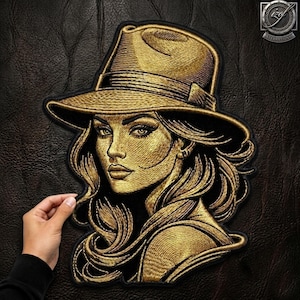 Pode incluir: Um emblema bordado a ouro com o retrato de uma pessoa usando um chapéu de aba larga. O design utiliza fio dourado metálico sobre um fundo preto para criar traços faciais detalhados, cabelo fluido e a textura do chapéu.