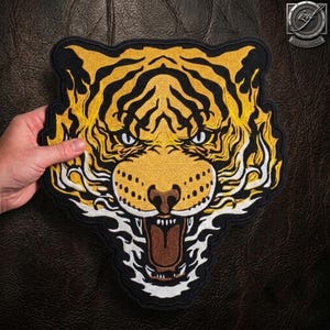 Op de afbeelding: Een geborduurde patch met een tijgerkop met gele vacht, zwarte strepen en witte vlamachtige accenten. De tijger heeft een open bek met zichtbare tanden en een bruine tong, omlijnd door een zwarte rand rondom de gehele vorm.