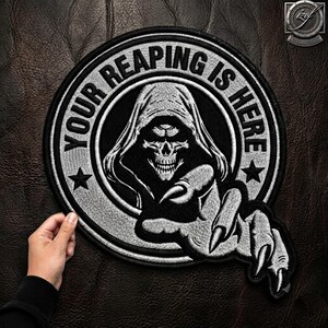 Könnte beinhalten: Ein runder bestickter Aufnäher mit einem schwarz-weißen Motiv einer Kapuzen-Totenkopffigur und einer ausgestreckten Skeletthand. Der Text "YOUR REAPING IS HERE" ist in Schwarz entlang des oberen Randes gestickt und von zwei kleinen Sternen flankiert.