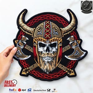 Op de afbeelding: Een ronde geborduurde patch met een schedel die een gouden gehoornde helm met baard draagt, geplaatst over twee gekruiste strijdbijlen. Het ontwerp bevat ingewikkelde rode knoopwerkpatronen op een zwarte achtergrond met gouden en zilveren accenten.