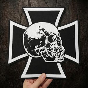 Può includere: Una toppa con teschio in bianco e nero con un design dettagliato del teschio centrato su una croce nera con bordi bianchi. La toppa è probabilmente per giacche o gilet.