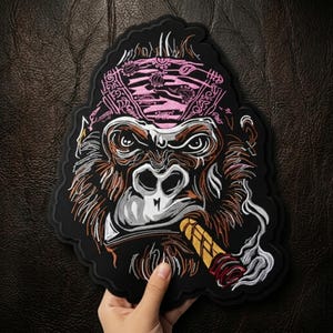 May include: Embroidered patch featuring a gorilla head wearing a pink bandana and smoking a cigar. The patch has a black border and detailed stitching in brown, white, and gold. The gorilla's expression is intense, with smoke rising from the cigar.