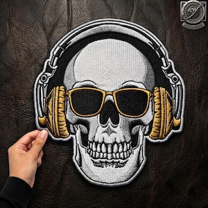 Puede incluir: Un parche bordado que muestra una calavera blanca con gafas de sol negras de montura dorada y auriculares grandes de color dorado. El diseño utiliza hilo negro, blanco y dorado para crear un gráfico detallado de una calavera con música.