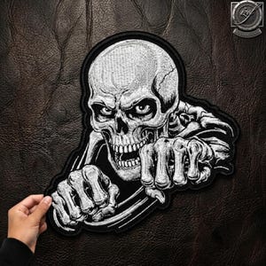 Könnte beinhalten: Ein gestickter Aufnäher mit einer detaillierten schwarz-weißen Illustration eines menschlichen Schädels, dessen skelettierte Hände ein Lenkrad umgreifen. Das Motiv ist in kontrastreichem weißem Garn auf schwarzem Grund gestickt.