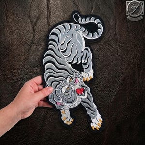 May include: An embroidered patch featuring a grey and white tiger with black stripes in a downward pouncing position. The tiger has a red mouth, pink nose, and yellow claws, outlined with a thick black border on a dark textured background.