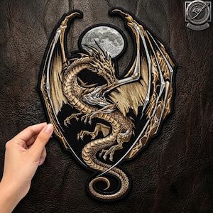 Pode incluir: Um emblema bordado apresentando um dragão dourado com detalhes prateados e asas abertas diante de uma lua cheia. O dragão possui uma longa cauda enrolada e detalhes intrincados de escamas, sobre um fundo de cor preta sólida.