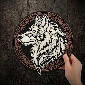 May include: A round black patch featuring a detailed white wolf head design. The wolf is surrounded by an intricate border with a brown outline. The patch is held against a dark brown leather-like surface.
