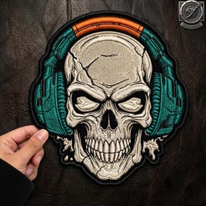 Peut inclure: Un écusson brodé représentant un crâne humain beige portant un casque audio bleu sarcelle avec un arceau orange. Le crâne présente des fissures visibles et des orbites profondes. L'écusson a une bordure noire et suit les contours du motif.