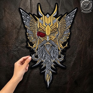 Op de afbeelding: Een geborduurde patch met een gestileerde bebaarde figuur met een gevleugelde helm in gouden, zilveren en zwarte draad. De figuur heeft één zichtbaar oog met een rode iris en een lange, gedetailleerde baard. Het ontwerp is symmetrisch met scherpe lijnen.