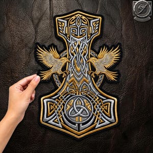 Könnte beinhalten: Ein bestickter Aufnäher mit einem zentralen Mjolnir-Hammer-Design aus gold- und silberfarbenem Garn. Der Hammer ist mit komplizierten Knotenmustern, zwei symmetrischen Raben mit ausgebreiteten Flügeln und einem Triquetra-Symbol verziert.