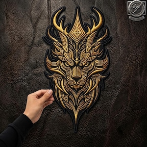 May include: A gold and black embroidered patch featuring a stylised lion head with intricate geometric patterns and horn-like protrusions. The design is symmetrical with sharp, pointed edges and a diamond-shaped detail at the top centre.