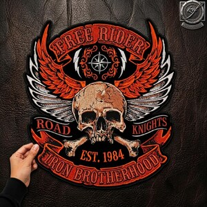 Könnte beinhalten: Ein großer gestickter Aufnäher mit einem zentralen menschlichen Totenkopf und gekreuzten Knochen. Über dem Schädel befinden sich orangefarbene Flügel und der Text FREE RIDER. Darunter steht ROAD KNIGHTS, EST. 1984 und IRON BROTHERHOOD auf orangefarbenen Bändern.