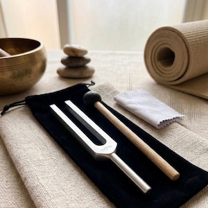 Könnte beinhalten: Eine silberne Stimmgabel mit der Aufschrift "432Hz" liegt auf einem schwarzen Samtbeutel, zusammen mit einem Holzschlägel und einem kleinen weißen Tuch. Eine gerollte beige Yogamatte, eine goldene Klangschale und gestapelte Steine sind ebenfalls zu sehen.