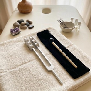 Peut inclure: Un diapason en métal argenté à trois branches lestées, marqué du texte OTTO 128Hz, accompagné d'un maillet en bois avec une tête en caoutchouc noir et d'une pochette de rangement en velours noir, le tout posé sur un tissu texturé blanc cassé.