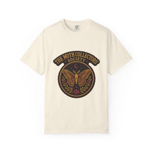 May include: A cream-coloured short-sleeve t-shirt featuring a circular graphic on the chest. The design displays a moth with intricate brown and gold patterns, surrounded by floral elements, a crescent moon, and the text THE MOTH COLLECTORS' SOCIETY.