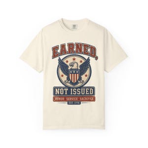 Pode incluir: Uma t-shirt de manga curta em tom off-white com um gráfico que apresenta uma águia-de-cabeça-branca, estrelas e riscas. O texto diz EARNED, NOT ISSUED, HONOR SERVICE SACRIFICE e EST. 1776. O design utiliza uma paleta de cores desgastada.