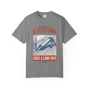 以下が含まれることがあります： グレーのTシャツで、コーキングガンが幅木にシーラントを塗布しているグラフィックイラストが描かれています。テキストは「Trade Wisdom No. 4, A LITTLE CAULK, GOES A LONG WAY」と書かれています。デザインには青と赤のインクが使用されています。