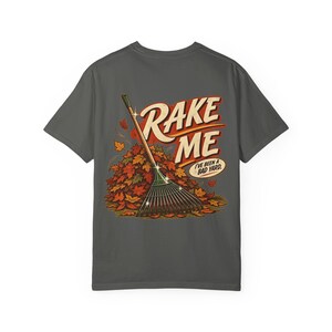 May include: A charcoal gray t-shirt featuring a graphic print of a garden rake resting against a large pile of colorful autumn leaves. The text RAKE ME is printed in large bold letters above a speech bubble that reads I'VE BEEN A BAD YARD.