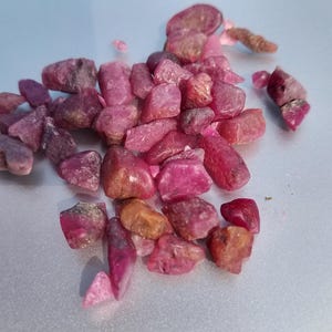 Può includere: Una collezione di gemme di rubino grezze e non tagliate. Le pietre variano per dimensioni e forma, mostrando sfumature di rosa e rosso con alcune tonalità marroni. Le gemme sono sparse su una superficie grigio chiaro.