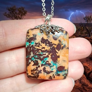 Peut inclure: Un collier avec une chaîne argentée et un pendentif rectangulaire. Le pendentif présente un motif unique de couleurs orange, marron et turquoise. Le pendentif est fixé à la chaîne par une bélière décorative en argent.