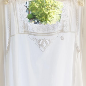 May include: A white sleeveless garment featuring a square neckline with intricate lace trim and scalloped edges. The chest area displays delicate white embroidery, including a small crown motif. The garment is suspended on a padded hanger.