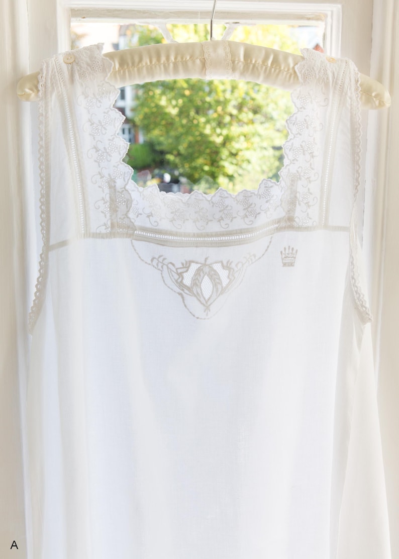 May include: A white sleeveless garment featuring a square neckline with intricate lace trim and scalloped edges. The chest area displays delicate white embroidery, including a small crown motif. The garment is suspended on a padded hanger.