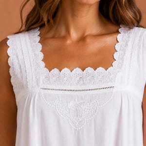 May include: A white sleeveless top featuring a square neckline with scalloped eyelet lace trim. The bodice includes intricate white floral embroidery centred below the neckline, complemented by vertical lace inserts along the shoulder straps.