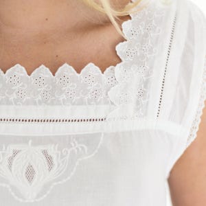 May include: A white sleeveless top featuring a square neckline with scalloped edges and intricate floral eyelet embroidery. The garment includes vertical pintuck pleats and a detailed decorative embroidered panel on the front chest area.