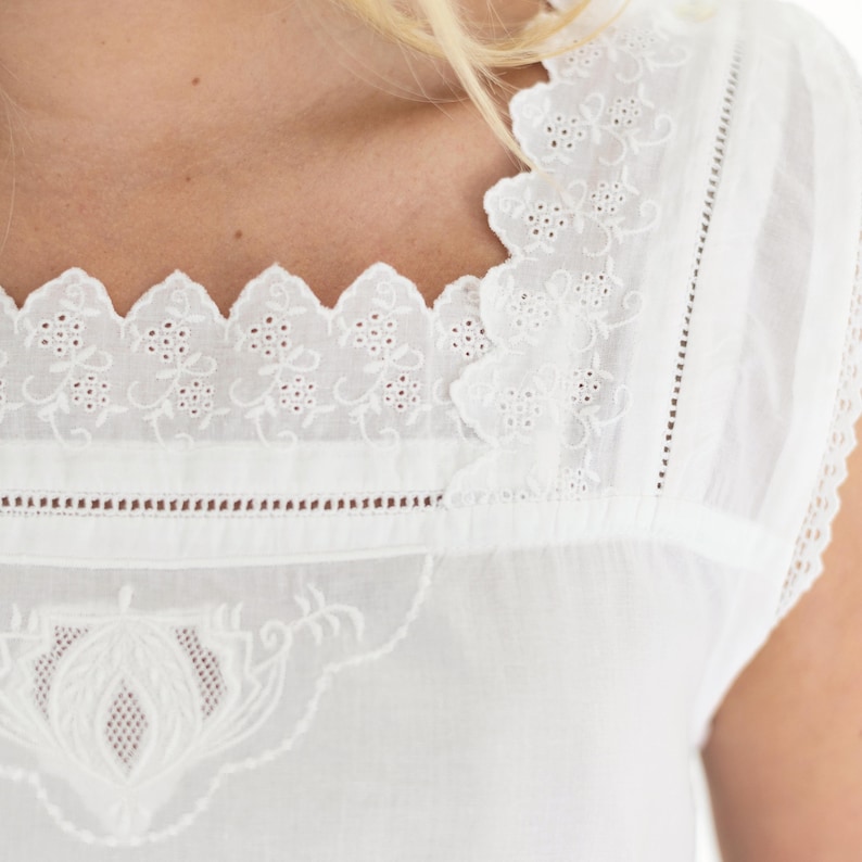 May include: A white sleeveless top featuring a square neckline with scalloped edges and intricate floral eyelet embroidery. The garment includes vertical pintuck pleats and a detailed decorative embroidered panel on the front chest area.