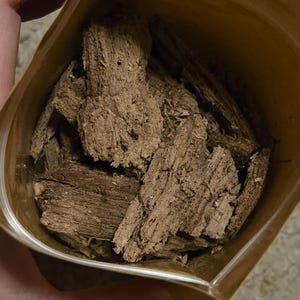 Pode incluir: Uma bolsa castanha aberta, cheia de vários pedaços de casca de madeira castanho-clara, rugosa e fibrosa. As peças têm uma textura irregular e serrilhada, apresentando veios naturais e bordas lascadas dentro do recipiente.
