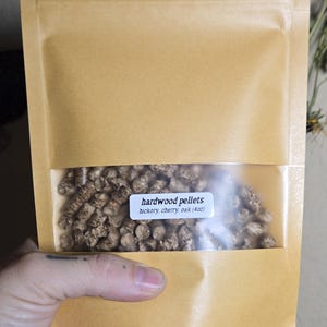May include: A tan paper pouch with a clear plastic window containing small brown hardwood pellets. A white label on the window reads: hardwood pellets, hickory, cherry, oak (4oz). The pouch is sealed at the top and has a flat bottom base.