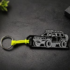 Peut inclure: Porte-clés SUV noir et argent avec un cordon jaune néon. Le porte-clés présente un contour détaillé d'un véhicule à quatre roues motrices. Le porte-clés est attaché à un anneau en métal argenté.