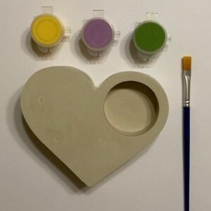 Può includere: Un kit di pittura in ceramica a forma di cuore con un'indentazione circolare. Include tre vasetti di vernice con vernice gialla, viola e verde e un pennello con manico blu. Il kit è progettato per progetti di pittura creativi.
