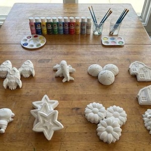 Op de afbeelding: Een verzameling ongeverfde keramische figuren, waaronder raketten, vliegtuigen, bollen, huizen, sterren, bloemen en bomen, op een houten tafel. Verfpotten, paletten en penselen suggereren een doe-het-zelf schilderactiviteit.