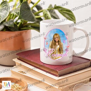 Puede incluir: Taza de cer&aacute;mica blanca con un retrato de una mujer con cabello rubio largo, mariposas rosas y el nombre "Hannah" en cursiva. La imagen est&aacute; sobre un fondo de cielo azul. La taza tiene un asa curva y est&aacute; sobre una pila de libros.