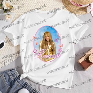 Puede incluir: Camiseta blanca con un retrato de una mujer con cabello rubio largo, mariposas y el nombre "Hannah" en escritura rosa. El retrato est&aacute; sobre un fondo de cielo azul dentro de un &oacute;valo rosa.