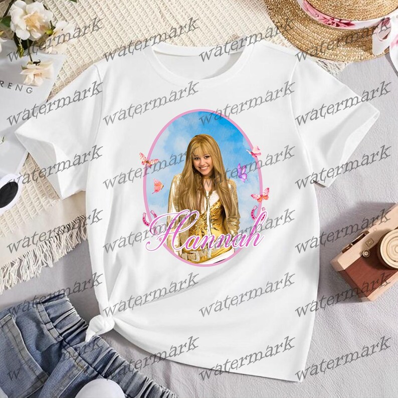 Puede incluir: Camiseta blanca con un retrato de una mujer con cabello rubio largo, mariposas y el nombre "Hannah" en escritura rosa. El retrato est&aacute; sobre un fondo de cielo azul dentro de un &oacute;valo rosa.