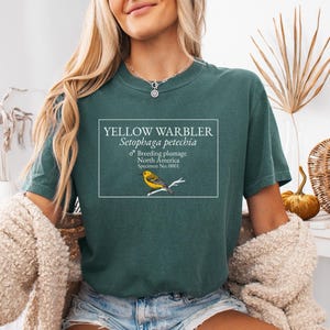 May include: A dark green short-sleeve t-shirt featuring a white rectangular graphic with the text YELLOW WARBLER Setophaga petechia, Breeding plumage, North America, Specimen No. 0001, and an illustration of a yellow bird perched on a branch.