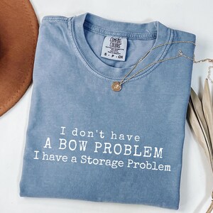 Op de afbeelding: Een blauw t-shirt met ronde hals en een vintage wassing. Het shirt is voorzien van witte tekst die luidt: I don't have A BOW PROBLEM I have a Storage Problem. Een goudkleurige ronde hangerketting rust op de halslijn van het shirt.
