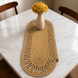 Pode incluir: Um caminho de mesa oval feito de corda trançada espessa em tom bronzeado dourado com um padrão decorativo de borda festonada. Um vaso de cerâmica liso de cor clara contendo um buquê de pequenas flores amarelas brilhantes está sobre ele.