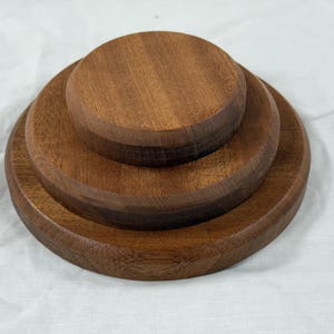May include: Three wooden, circular trays of varying sizes stacked together. The wood has a warm, brown tone with visible grain. The trays are arranged in a tiered fashion, with the largest at the base and the smallest on top. The background is a white, textured surface.