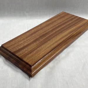 Può includere: Una base espositiva rettangolare in legno con una ricca finitura marrone scuro. La base presenta un design a strati con un bordo leggermente rialzato, che mette in risalto le venature naturali del legno. Adatta per oggetti da collezione.