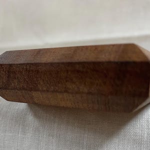 May include: A rectangular wooden object with a faceted design. The wood has a warm, brown tone and a visible grain. The object is set against a white, textured background.